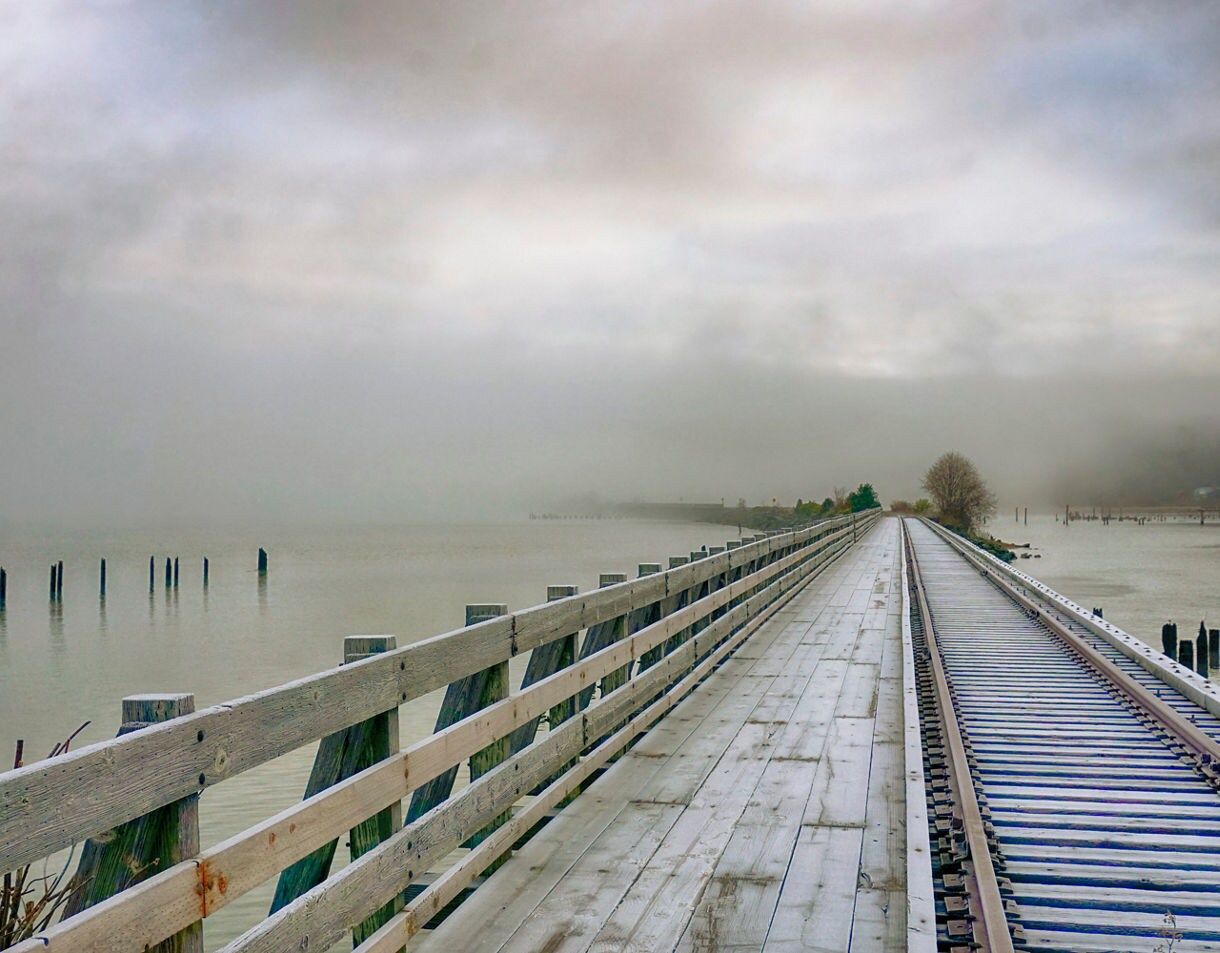 Long wooden boardwalk with adjacent train tracks leading into thick coastal fog, calm water on both sides and scattered pilings rising from the inlet.