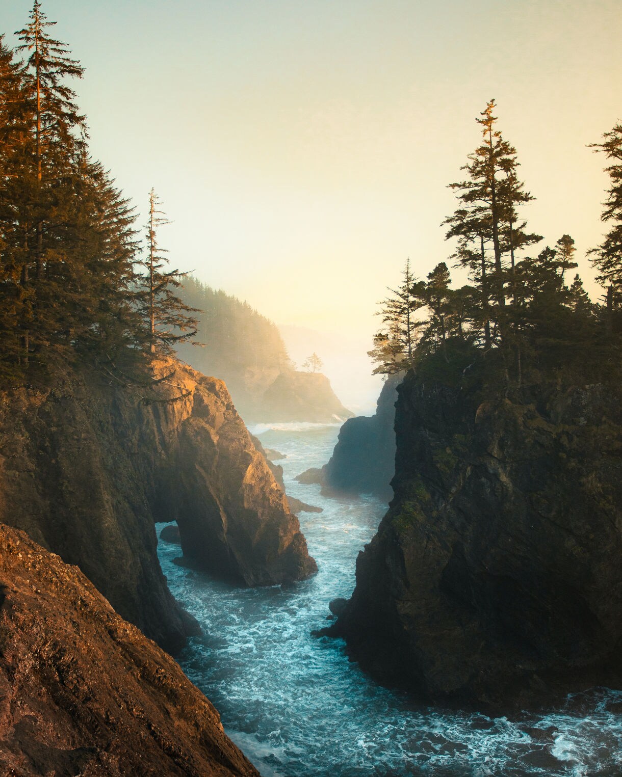 Narrow inlet framed by steep rocky cliffs topped with evergreens, with waves crashing below and soft sunset haze drifting through distant forested hills.