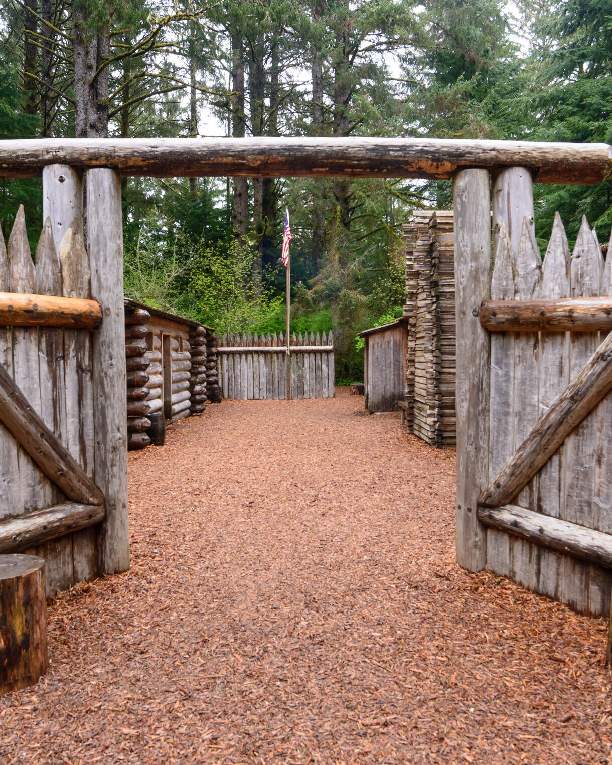 Entrance to Fort Clatsop with open log gates leading into a courtyard surrounded by wooden cabins, set amid dense forest and a flagpole in the center.