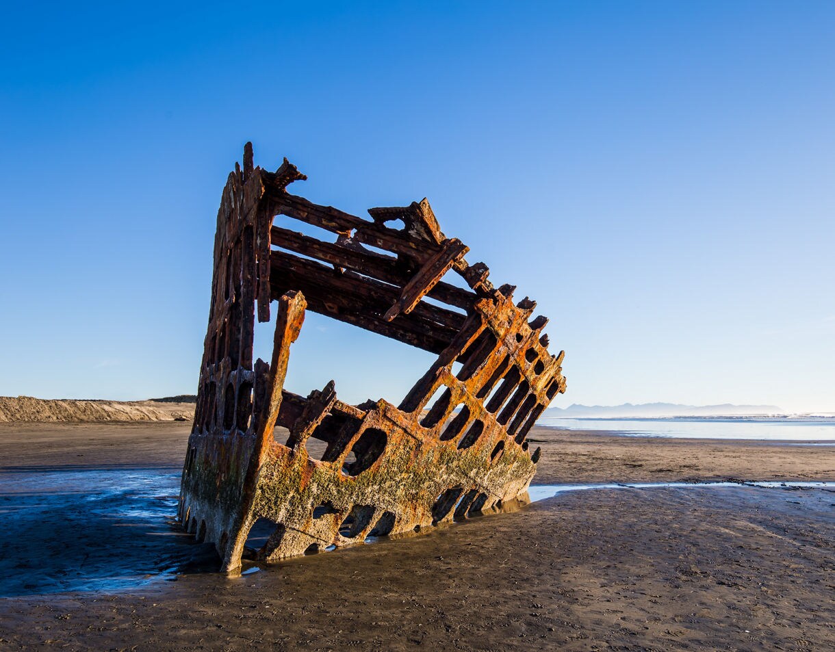 Close-up of the Peter Iredale shipwreck at Fort Stevens, showing its corroded steel ribs angled in the sand with a wide, empty beach and calm ocean in the background.