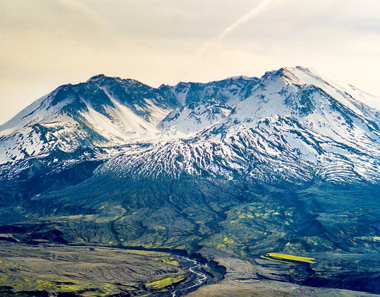 Expansive view of Mount St. Helens showing its snow-covered crater, dark volcanic slopes and a winding river cutting through the barren landscape below.