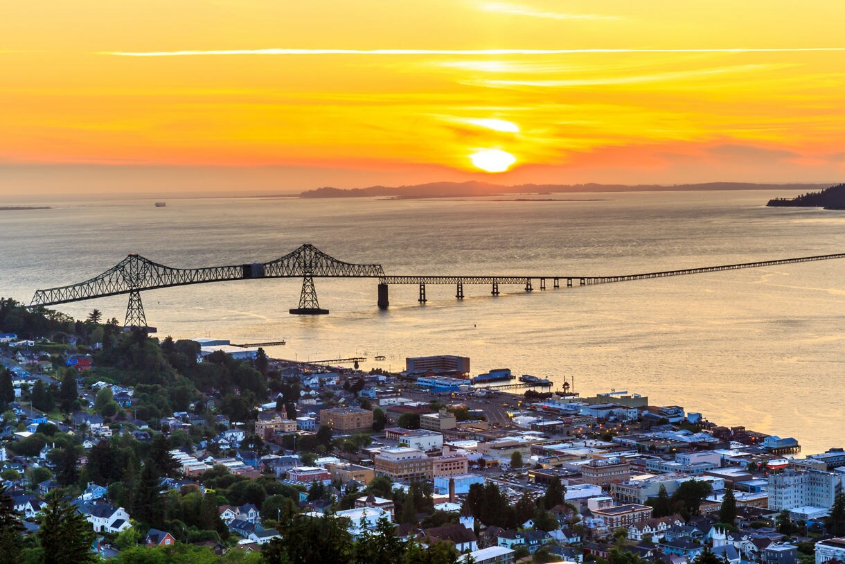 Aerial view of Astoria at sunset with the Astoria-Megler Bridge spanning the Columbia River, warm golden light reflecting on the water and the town’s buildings in the foreground.