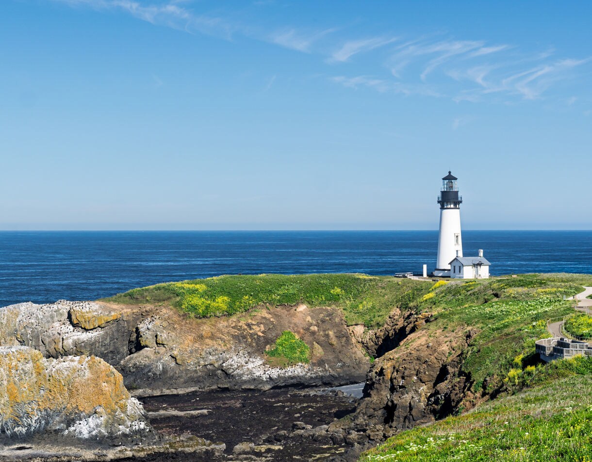 View of Yaquina Head Lighthouse perched on a grassy cliff overlooking the deep blue ocean, with rocky outcrops and bright green vegetation in the foreground.