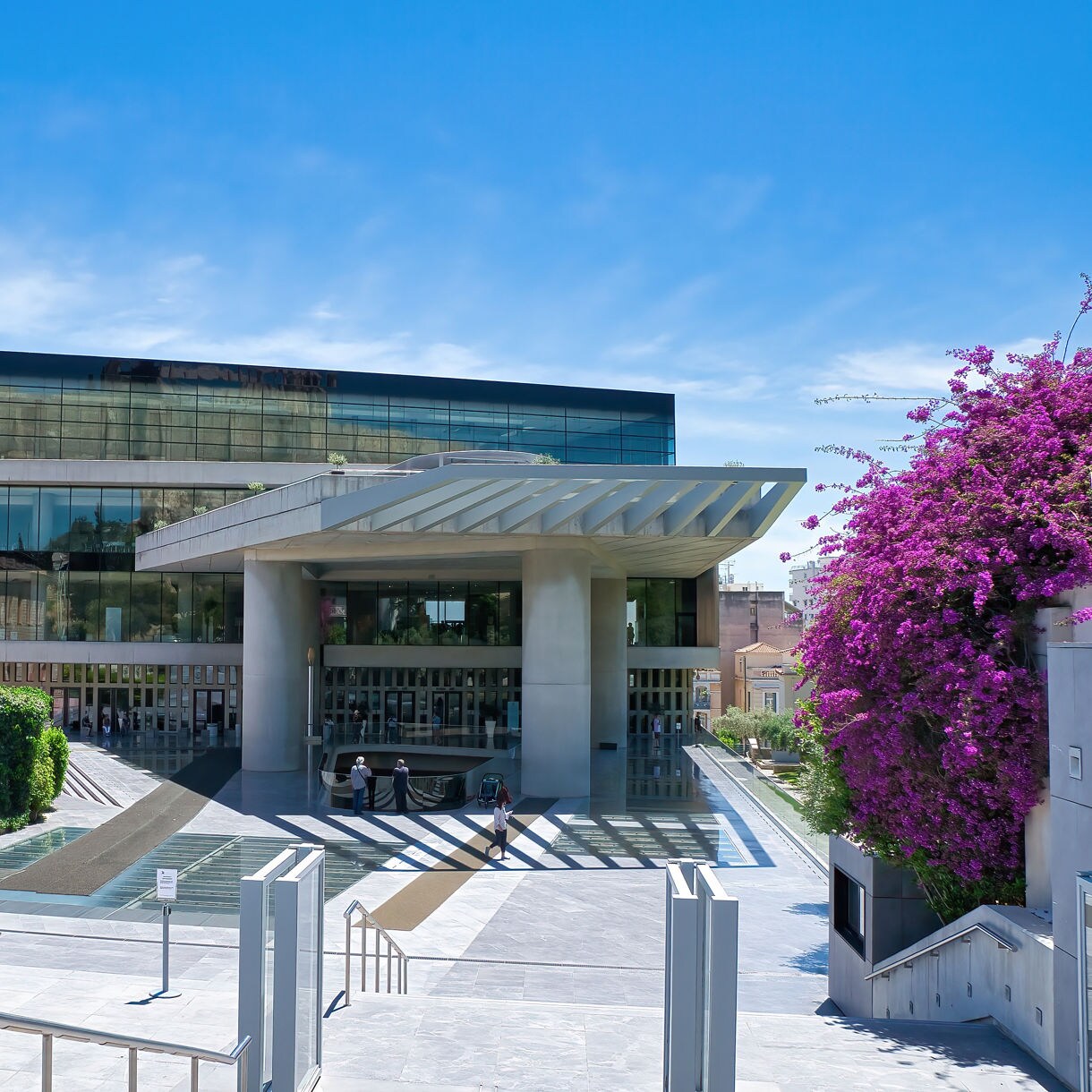 Exterior view of the Acropolis Museum in Athens featuring a modern glass-fronted building with a wide entrance, alongside a historic stone structure and landscaped gardens.
