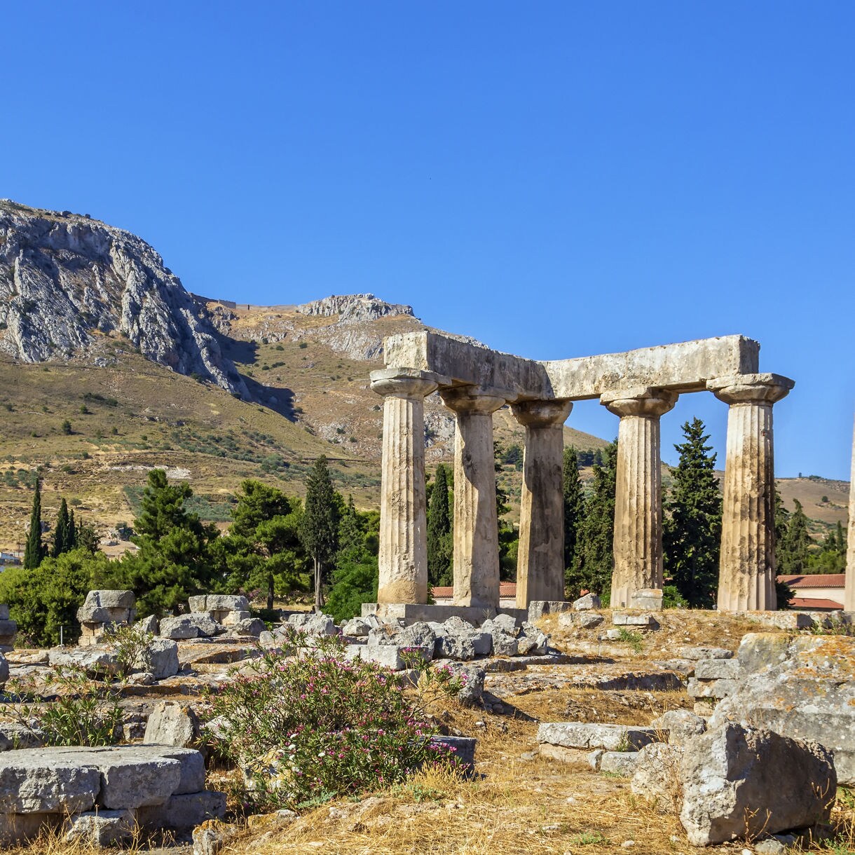 Ruins of the Temple of Apollo in Corinth with several standing stone columns, rocky remains scattered around and a steep green mountain rising in the background.