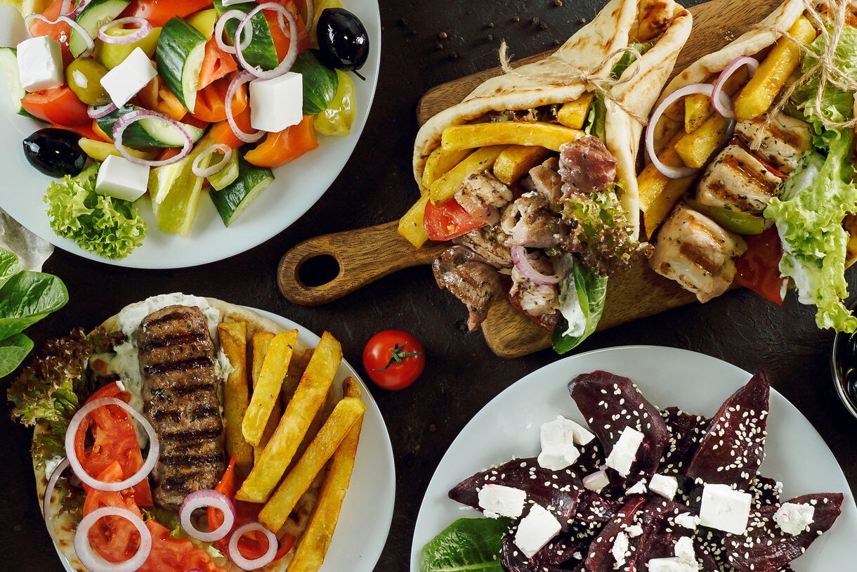 Overhead view of a Greek spread featuring grilled chicken and pork gyros in pita, fries, halloumi, Greek salad with feta, beet salad and assorted plates arranged on a dark table.
