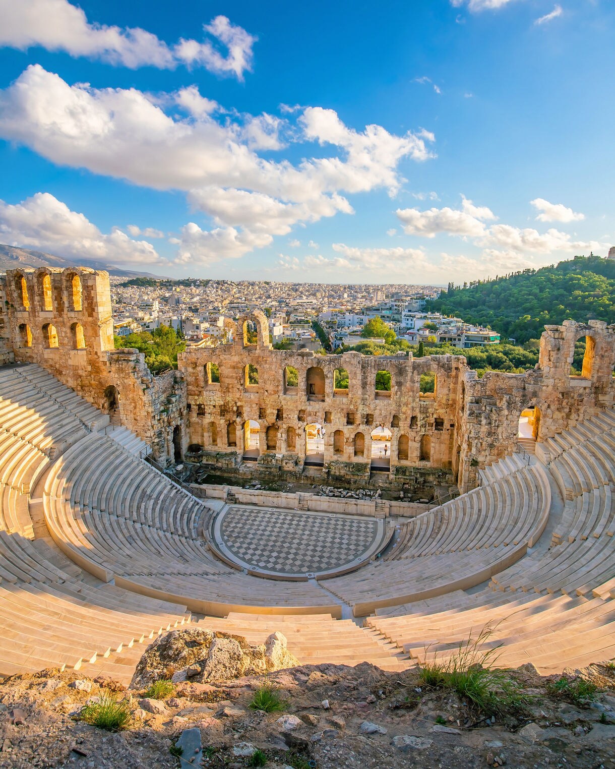 The Odeon of Herodes Atticus in Athens with semicircular marble seating, a tall stone backdrop and views of the city and green hills beyond under a partly cloudy sky.