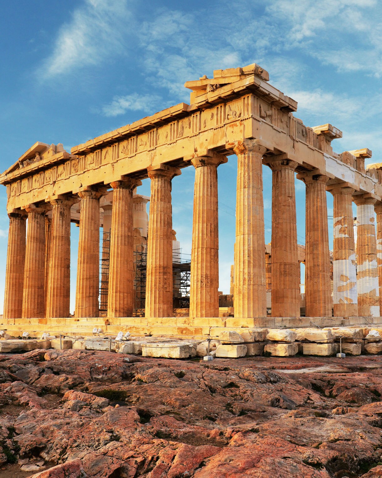 The Parthenon, an iconic ancient Greek temple, stands atop the Acropolis in Athens, Greece. The structure features classical Doric columns and is surrounded by rocky terrain under a bright blue sky. This historic site is a symbol of ancient Greek architecture and culture.
