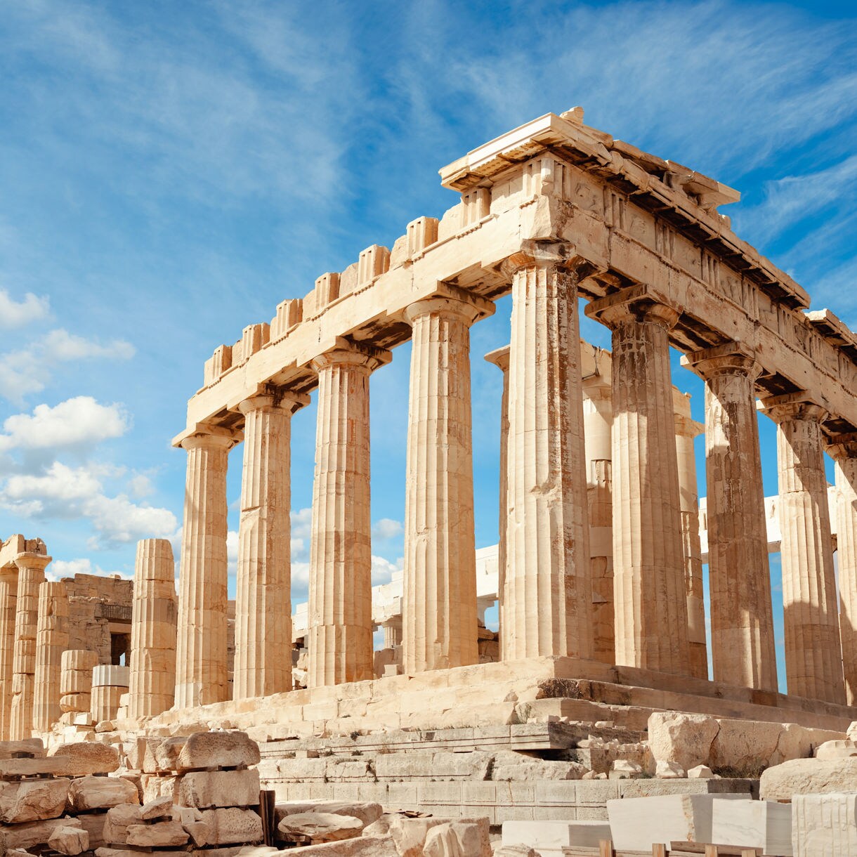 The Parthenon on the Acropolis in Athens with tall Doric columns and stone ruins in the foreground under a blue sky with scattered clouds.