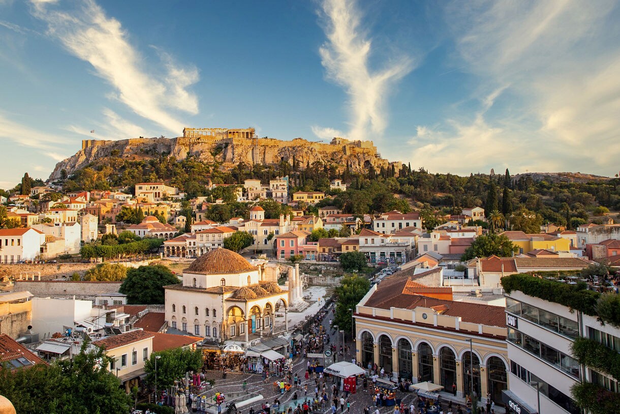 View of Monastiraki Square filled with shops and crowds with the Acropolis and Parthenon rising above the city at golden hour.