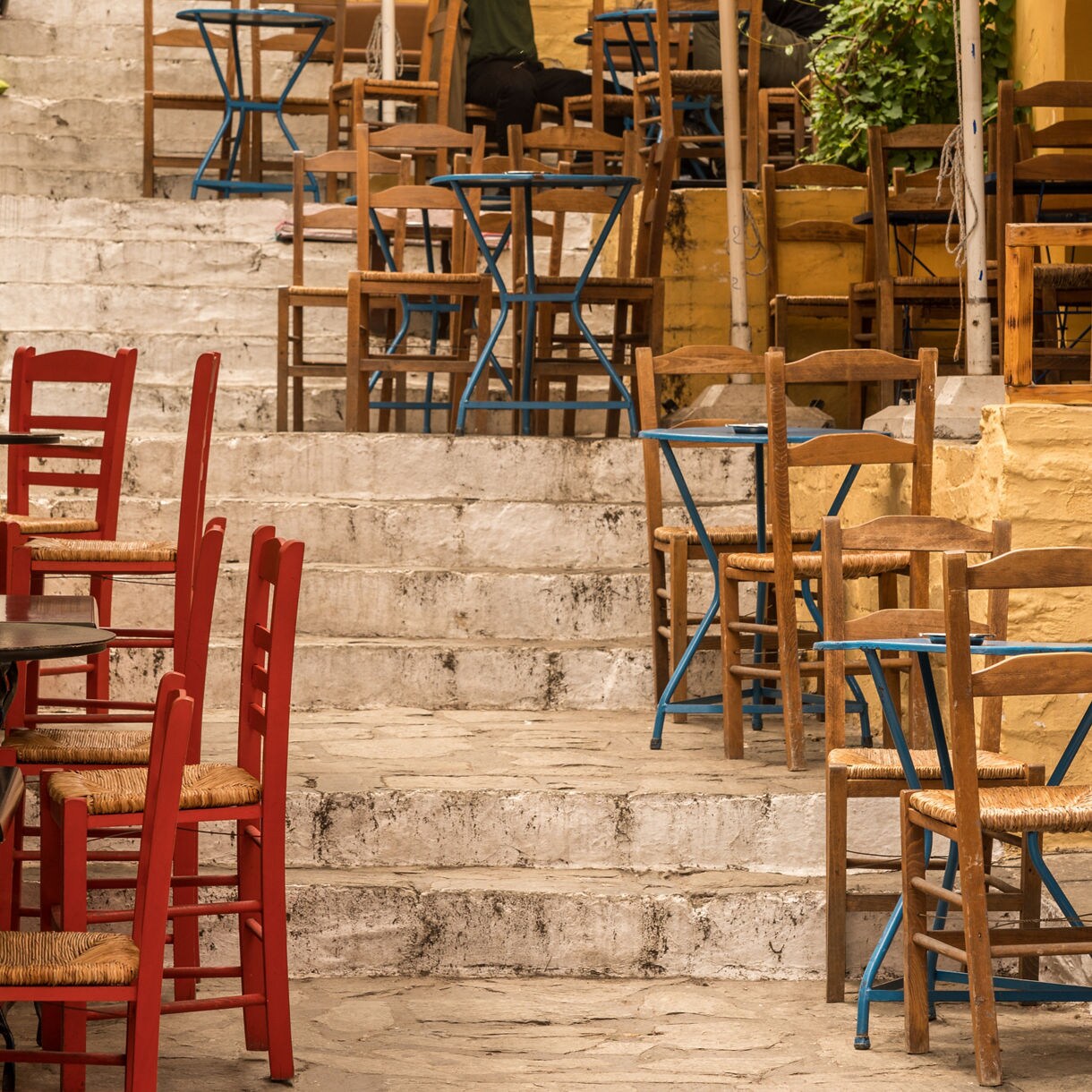 Outdoor seating on stone steps in Athens with colorful wooden chairs and small café tables arranged against yellow-painted walls.