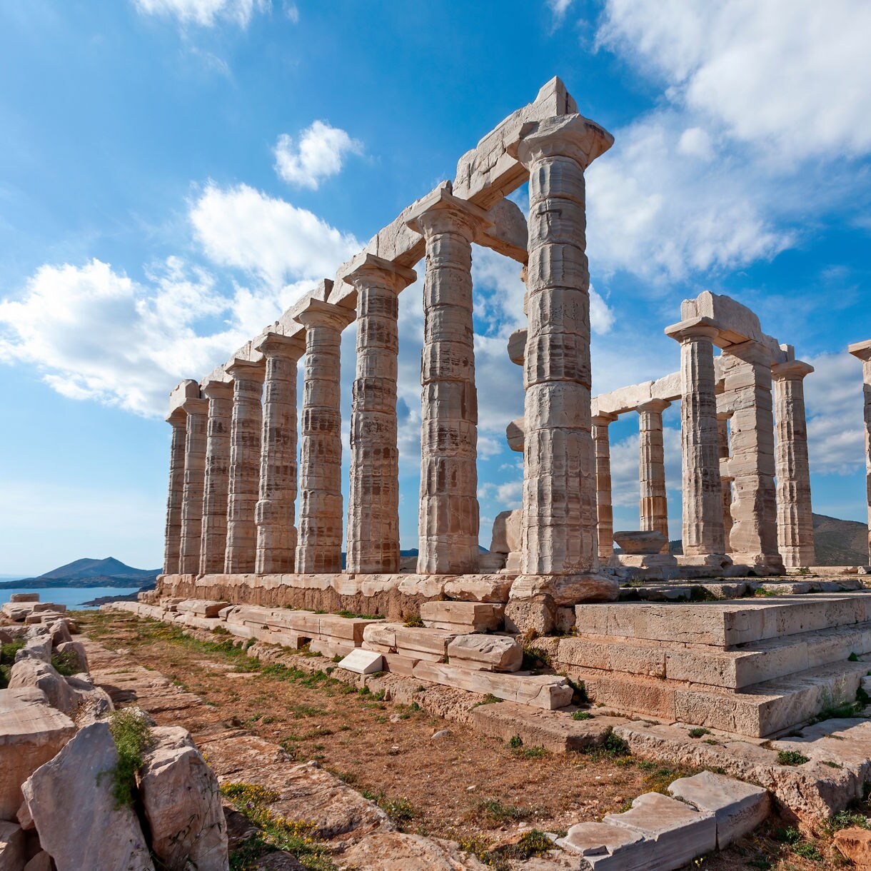 The Temple of Poseidon at Cape Sounion in Greece with tall marble columns standing on a rocky hilltop overlooking the deep blue Aegean Sea and a distant island.