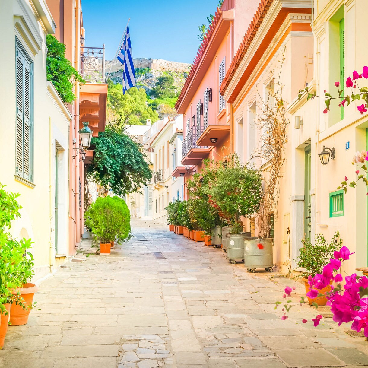 A narrow street in Athens’ Plaka neighborhood lined with pastel-colored houses, potted plants and bright pink bougainvillea under a clear blue sky.