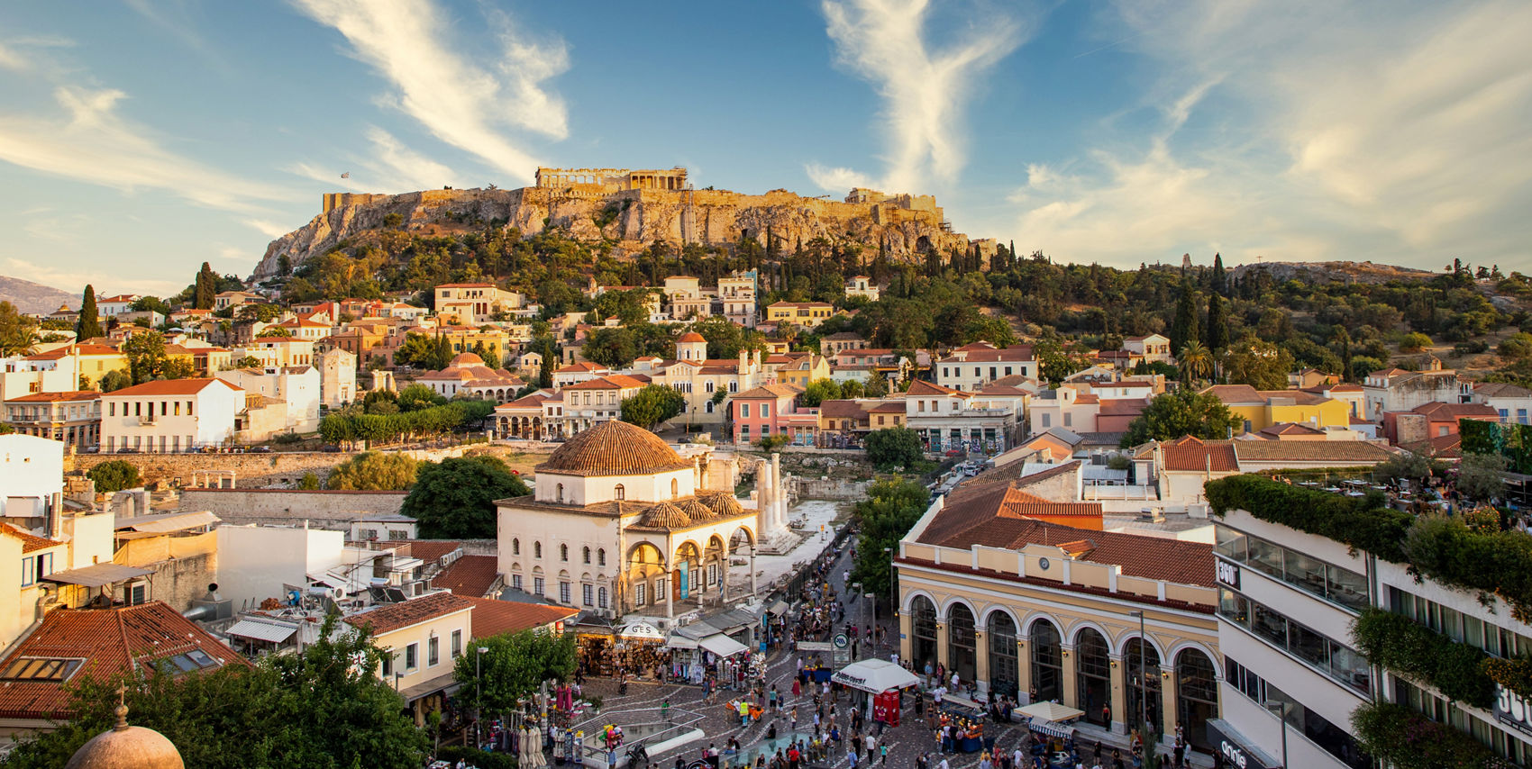Elevated view of Athens showing colorful neighborhoods and bustling streets beneath the Acropolis, with ancient ruins crowning the rocky hill under a dramatic sky.