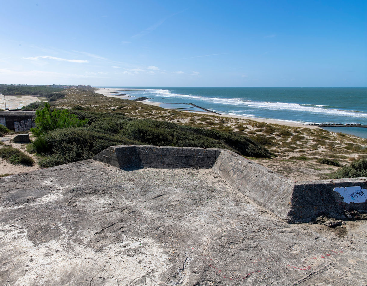 A weathered concrete WWII-era bunker sits on sandy dunes overlooking a wide coastline with rolling waves and low vegetation under a bright blue sky. 
