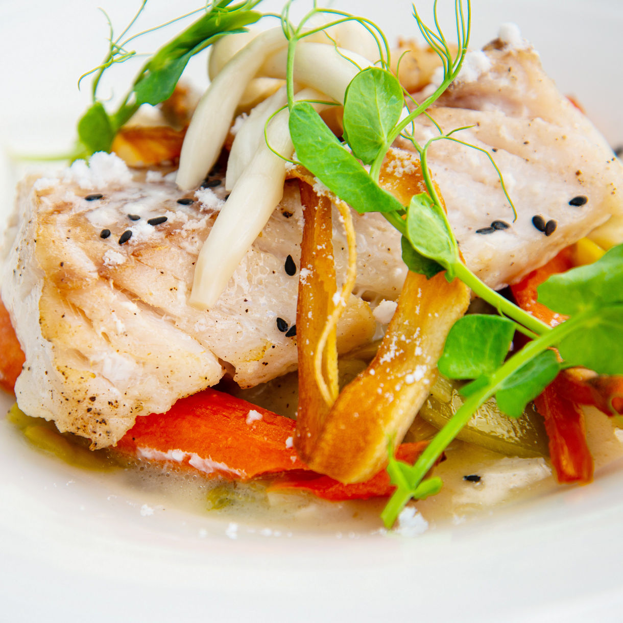 A plated seared fish fillet garnished with pea shoots and enoki mushrooms, served over roasted carrots and vegetables in a light broth.