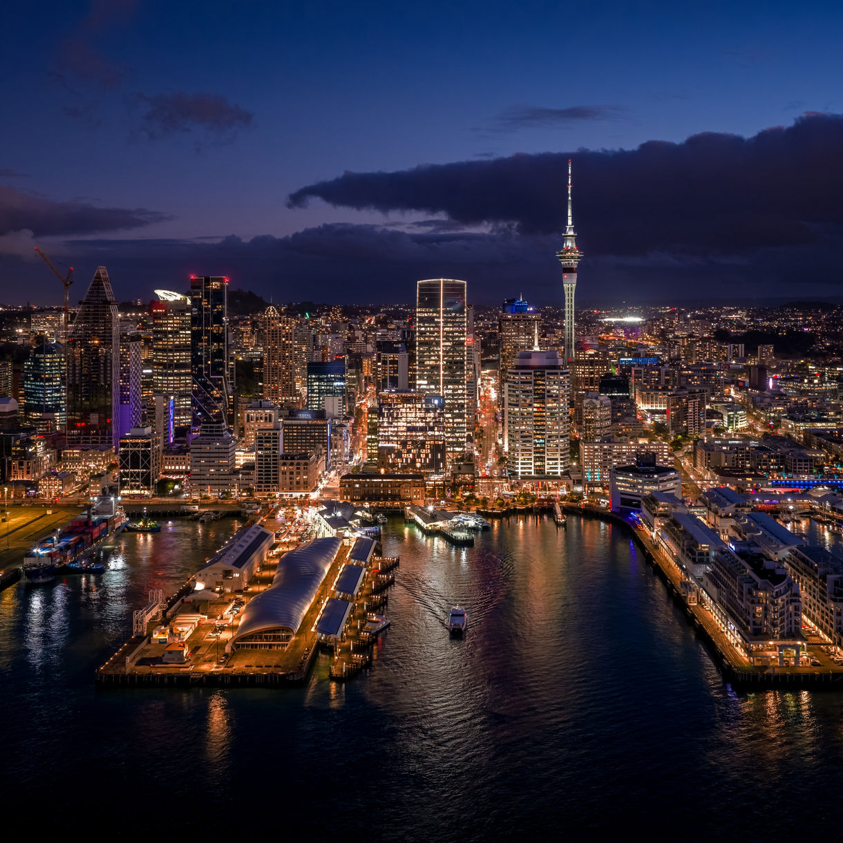 Nighttime aerial view of Auckland's illuminated skyline and Sky Tower, with a busy waterfront harbor and piers in the foreground.