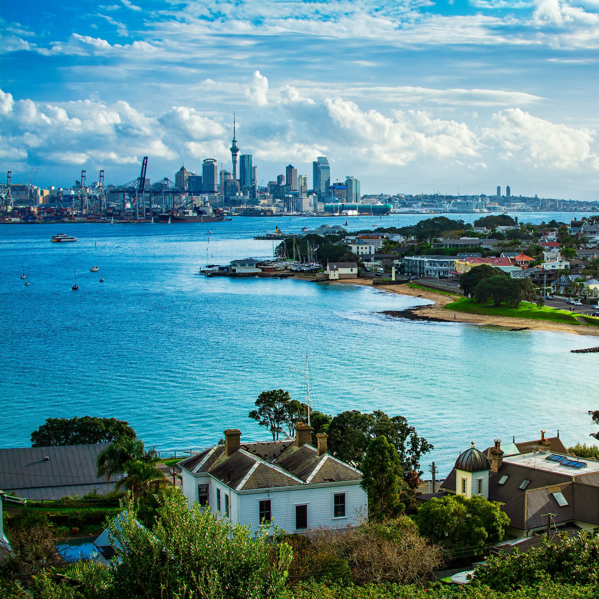A curved sandy beach and waterfront neighborhood in the foreground with turquoise harbor water leading toward Auckland's city skyline and port cranes under a partly cloudy sky.