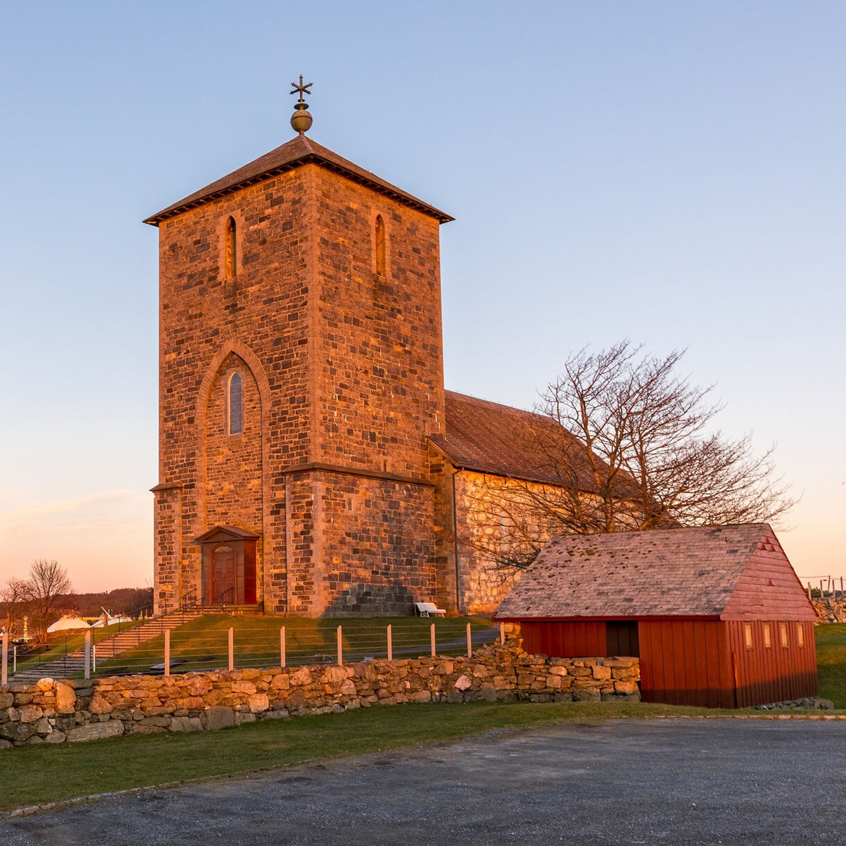 Avaldsnes Stone Church in Norway, built from grey stone with a tall square tower, glows in warm evening light beside a stone wall and small red outbuilding overlooking the water.