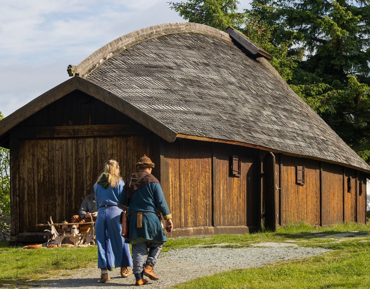 Two people dressed in Viking-era clothing approach a wooden longhouse with a curved roof at Avaldsnes Viking Farm, surrounded by trees and grassy fields.