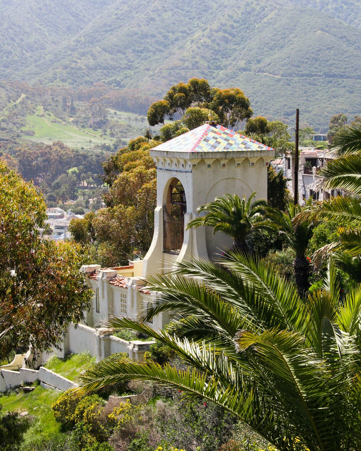 A white Spanish-style tower with a multicolored tiled roof rises among palm trees, flowering shrubs and lush green hills above Avalon.
