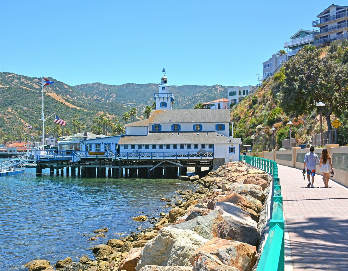  A couple walks along a paved waterfront path beside rocky shores and a blue wooden building on stilts in Avalon on Catalina Island, with green hills rising in the background.