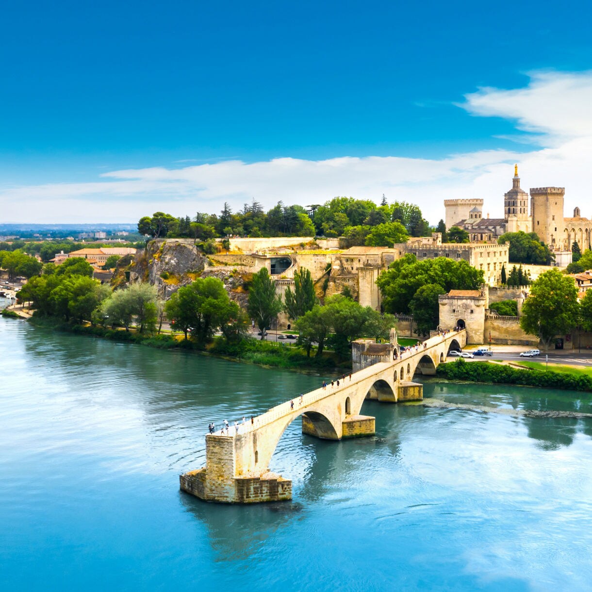 Aerial view of Avignon, France, showing the medieval Pont Saint-Bénézet stretching into the Rhône River with the Palais des Papes and historic city buildings in the background.