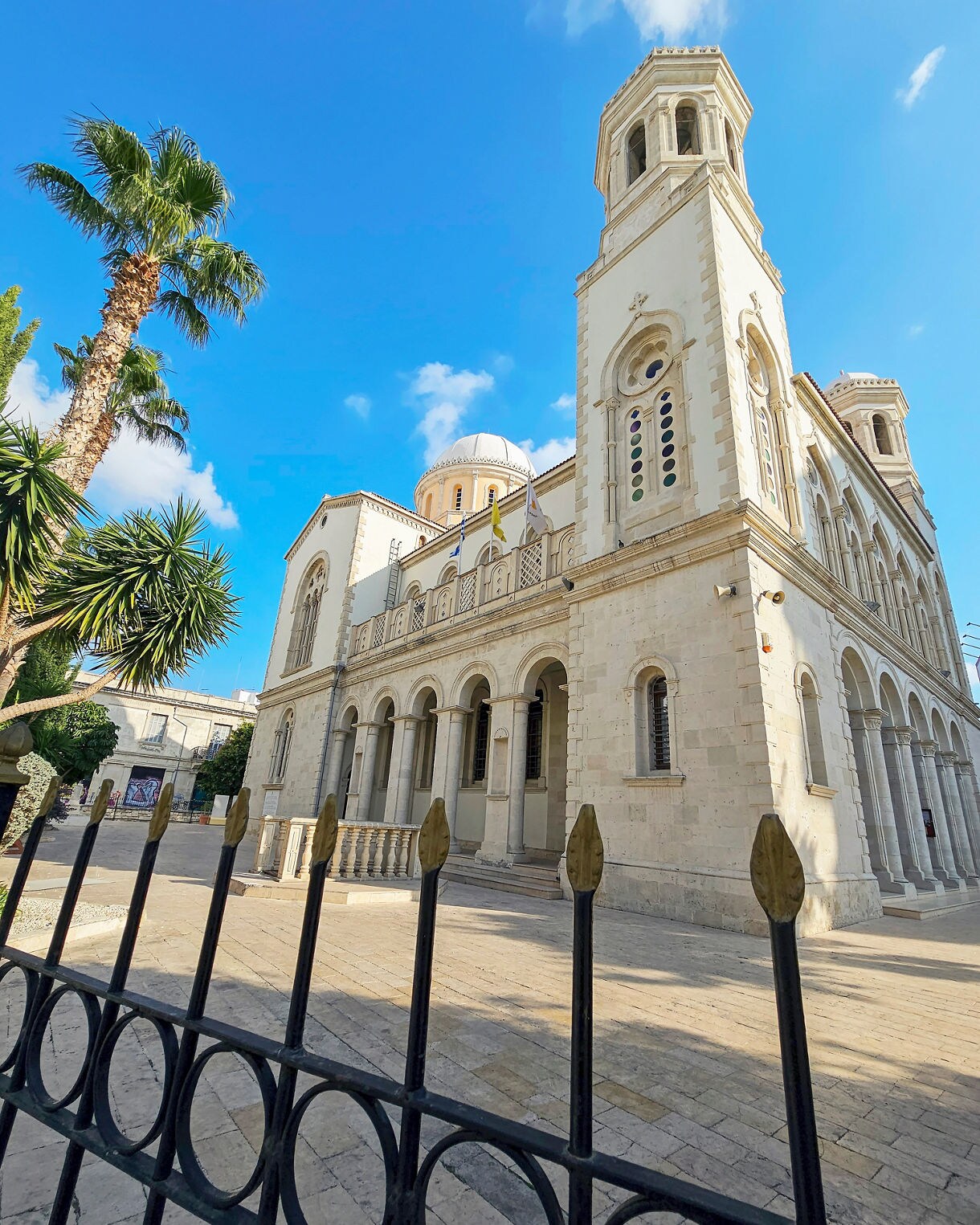 Exterior view of Ayia Napa Cathedral in Limassol, showing its tall white stone towers, arched entrance, domed roof and surrounding palm trees under a bright blue sky, with a black iron fence in the foreground.