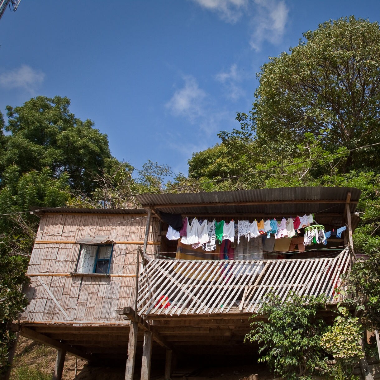 Rustic bamboo house on stilts with a corrugated metal roof and laundry hanging across the balcony, surrounded by dense green trees under a bright blue sky.