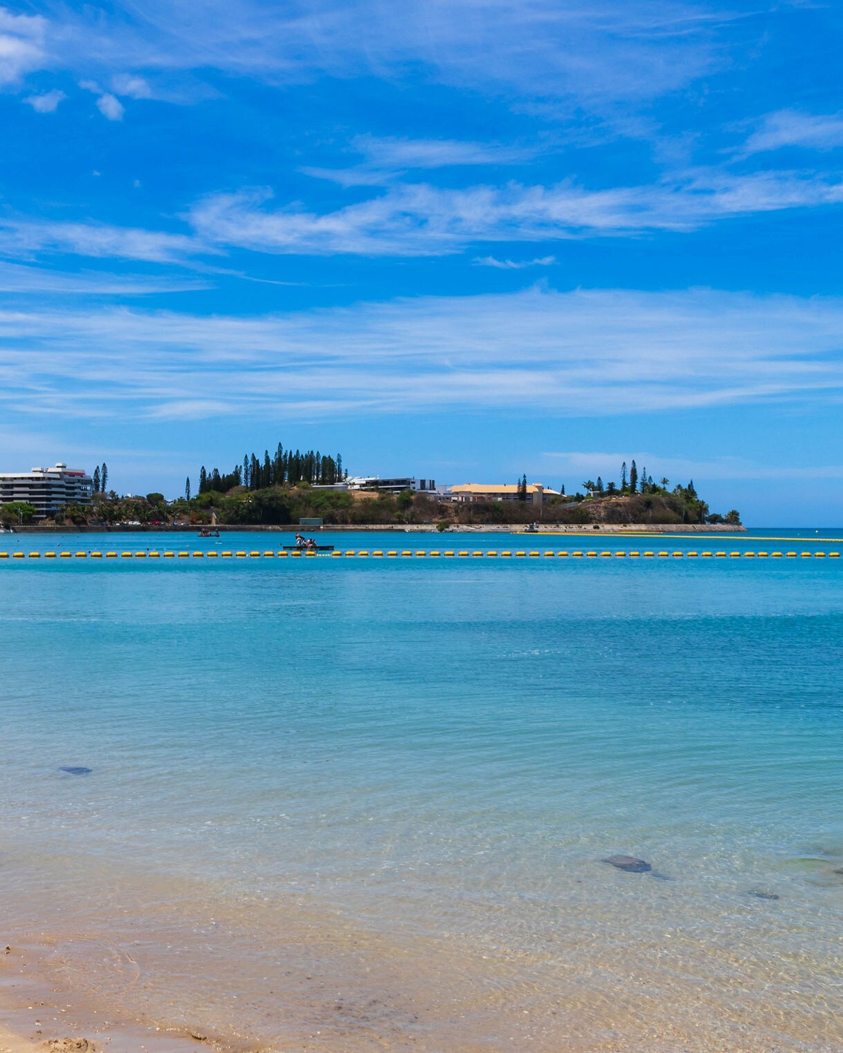 View of Baie des Citrons with gentle turquoise water, people swimming near shore and hotel buildings along the coastline under a blue sky.