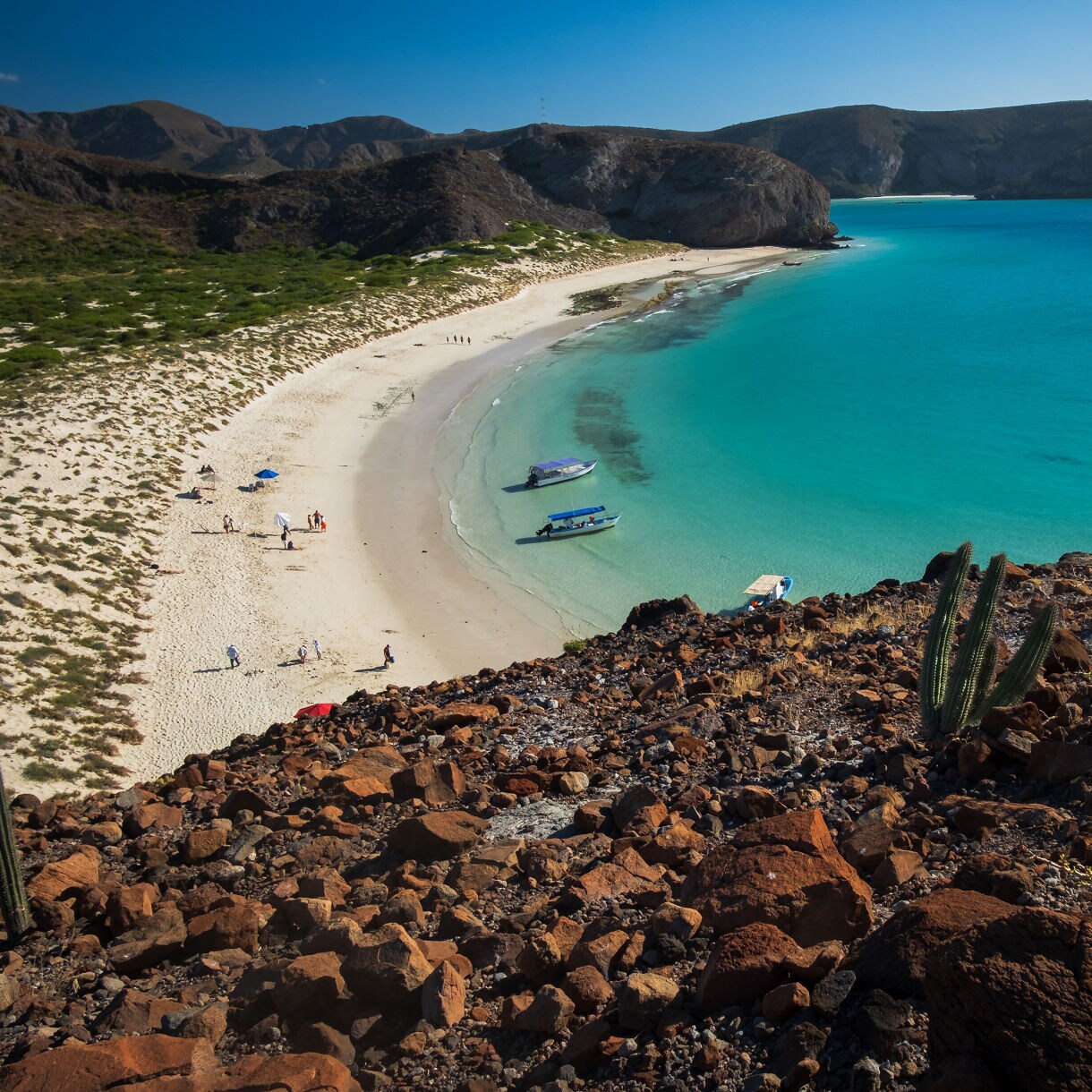 Aerial view of Balandra Beach in La Paz, Mexico, showing turquoise water, sandy shore, anchored boats and rocky desert hills with cacti.