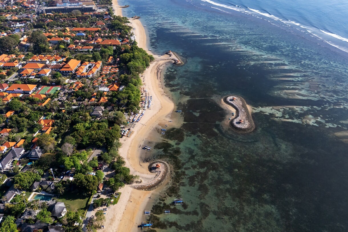 Aerial view of Bali’s Benoa Peninsula showing a long sandy shoreline lined with orange-roofed buildings, lush trees, and curved stone breakwaters extending into shallow turquoise water with visible reefs.