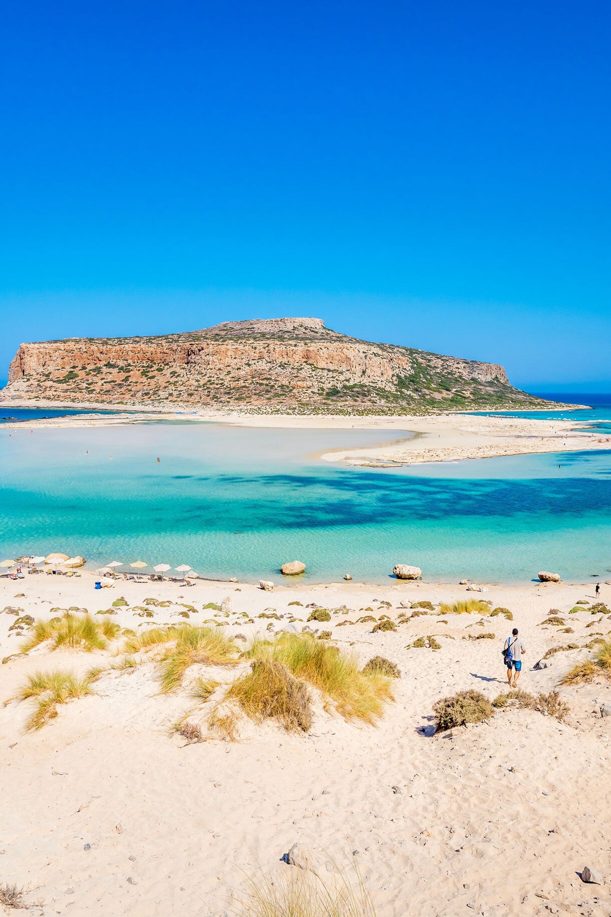 View of Balos Lagoon in Crete with shallow turquoise water, sandy beaches and a rocky island rising in the background under a clear blue sky.