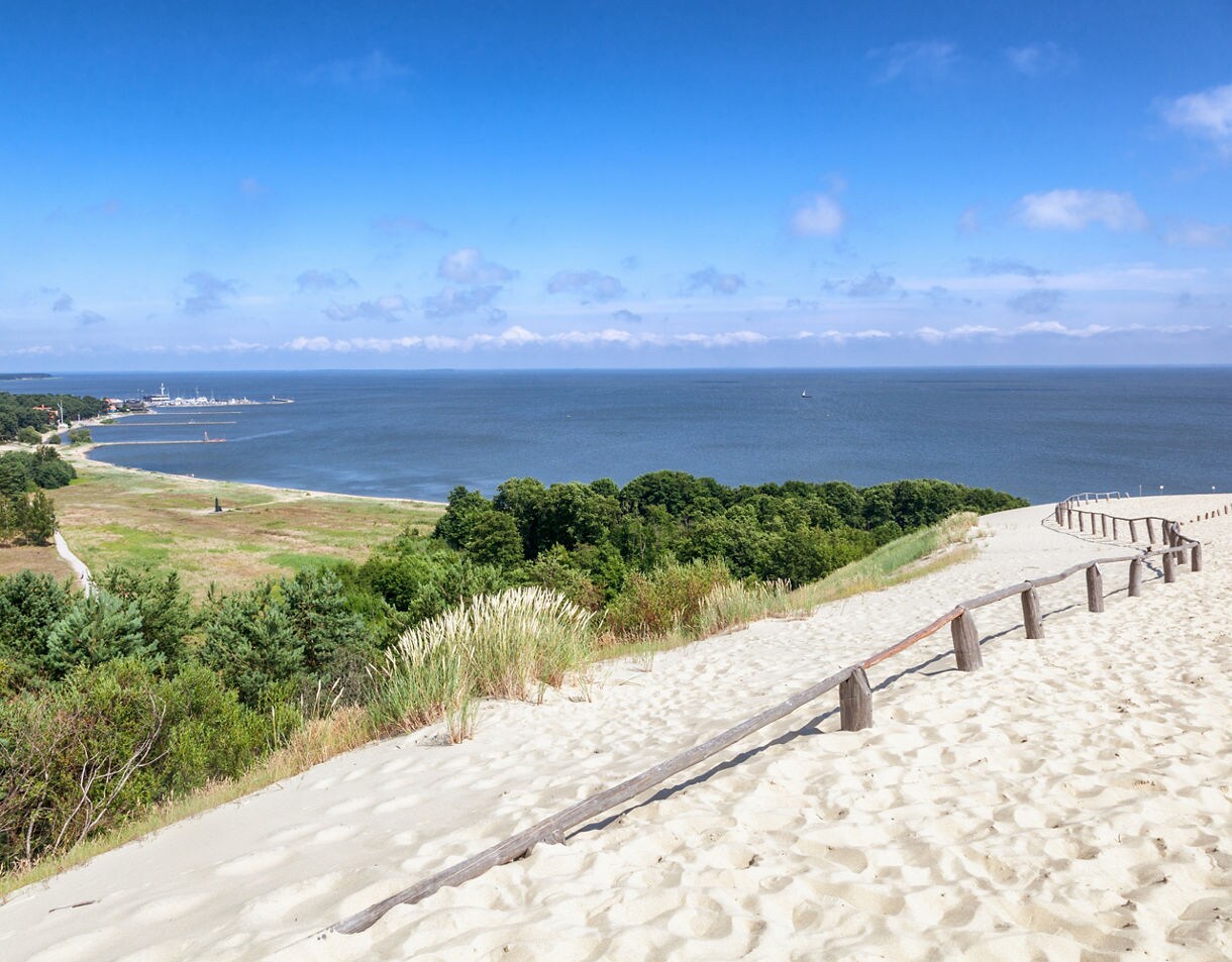 Expansive sand dunes overlooking the Baltic Sea with grassy patches, forest and a distant port in view.