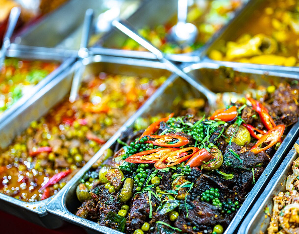 Trays of colorful Thai street food curries topped with fresh chilies, herbs and green peppercorns at a market stall.