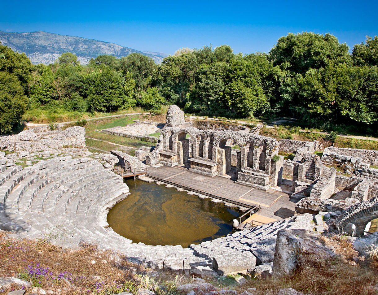 Ruins of the Baptistery at Butrint featuring stone archways, a semicircular theater-like structure and a shallow water-filled center surrounded by dense green forest and distant mountains.