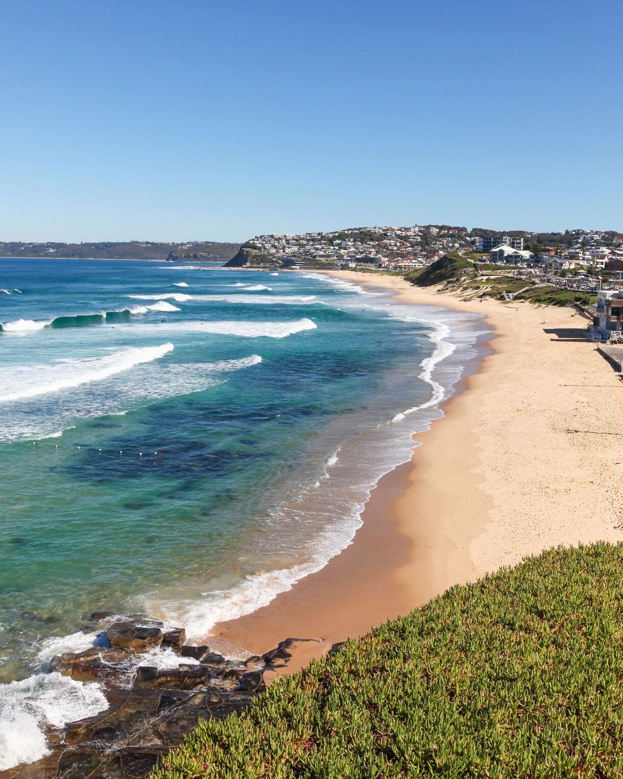 View of Bar Beach with clear turquoise waves, pale golden sand and coastal houses on low hills under a bright blue sky.