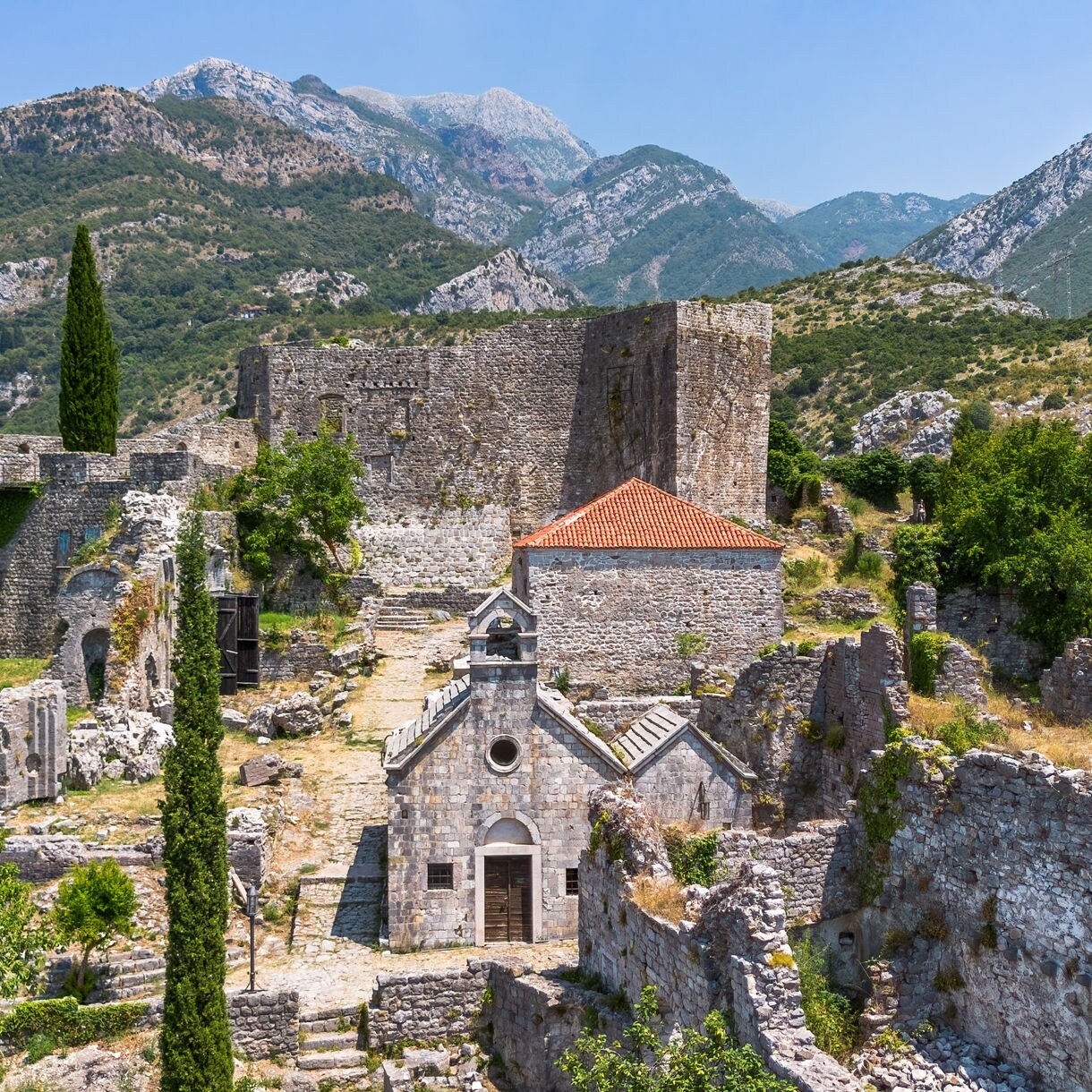 Ruined stone buildings and a small chapel surrounded by greenery in Stari Bar, Montenegro, with rugged mountains in the background.