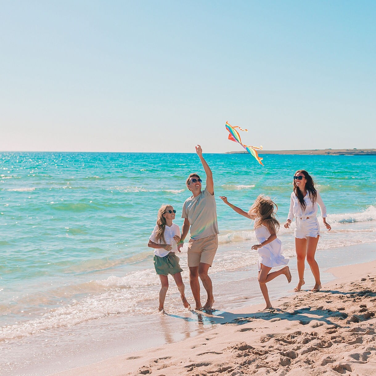 A family of four enjoys a bright, sunny day at the beach, flying a colorful kite near the shoreline. The group includes two adults and two children, all dressed in casual summer clothing. The turquoise water and clear sky create a cheerful, relaxed atmosphere. The scene captures playful movement and togetherness in a natural coastal setting.