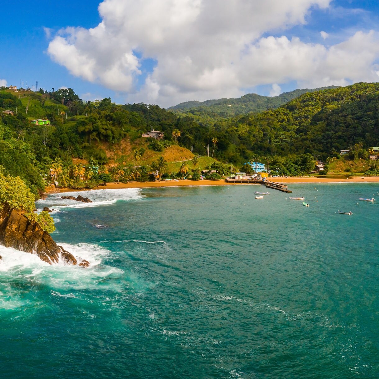 Panoramic view of a Caribbean coastline with green hills, rocky headlands and a calm bay dotted with small boats and turquoise ocean water.