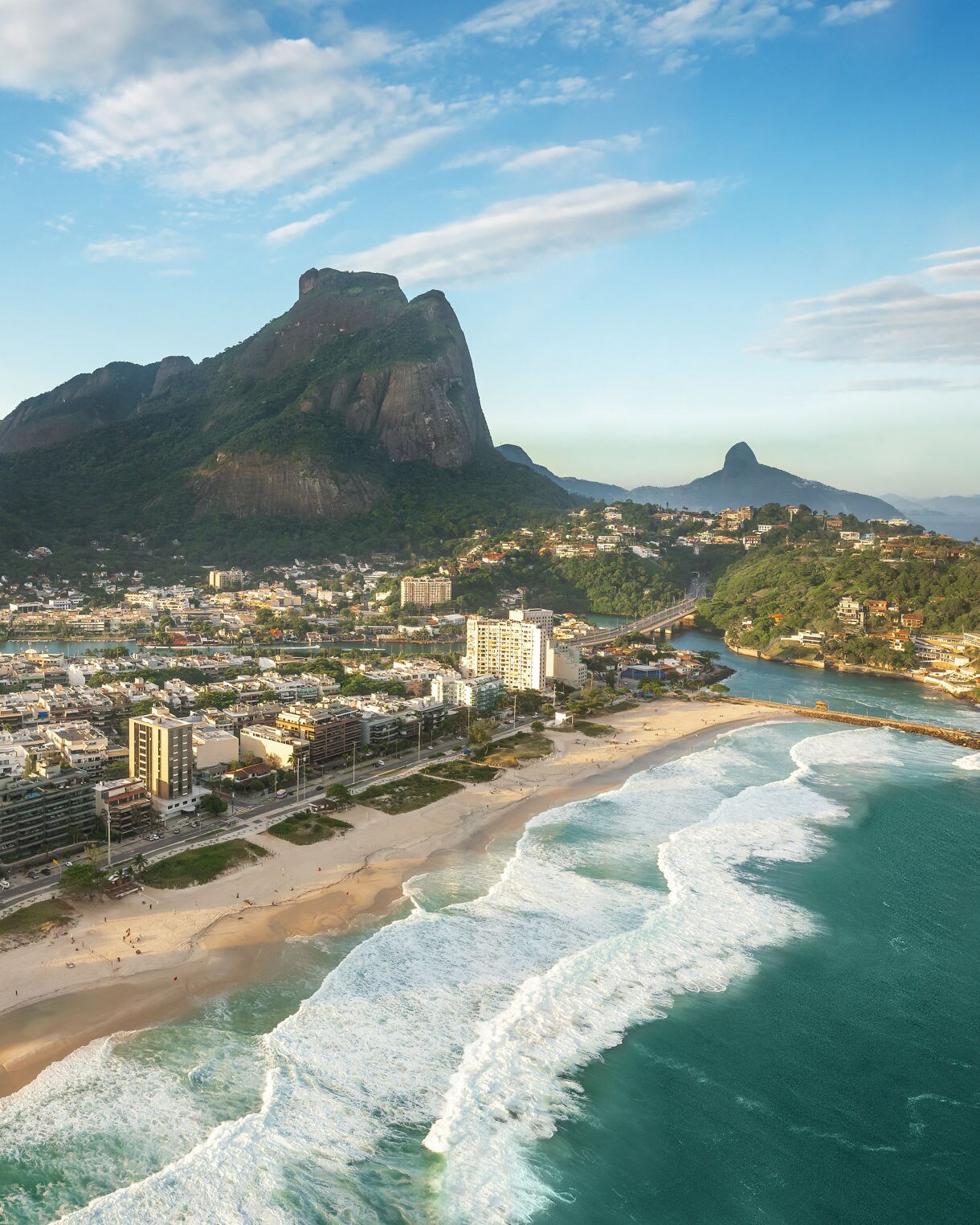 Aerial view of Barra da Tijuca showing wide sandy beaches, turquoise surf, modern beachfront buildings and lush green mountains rising behind the neighborhood under soft evening light.