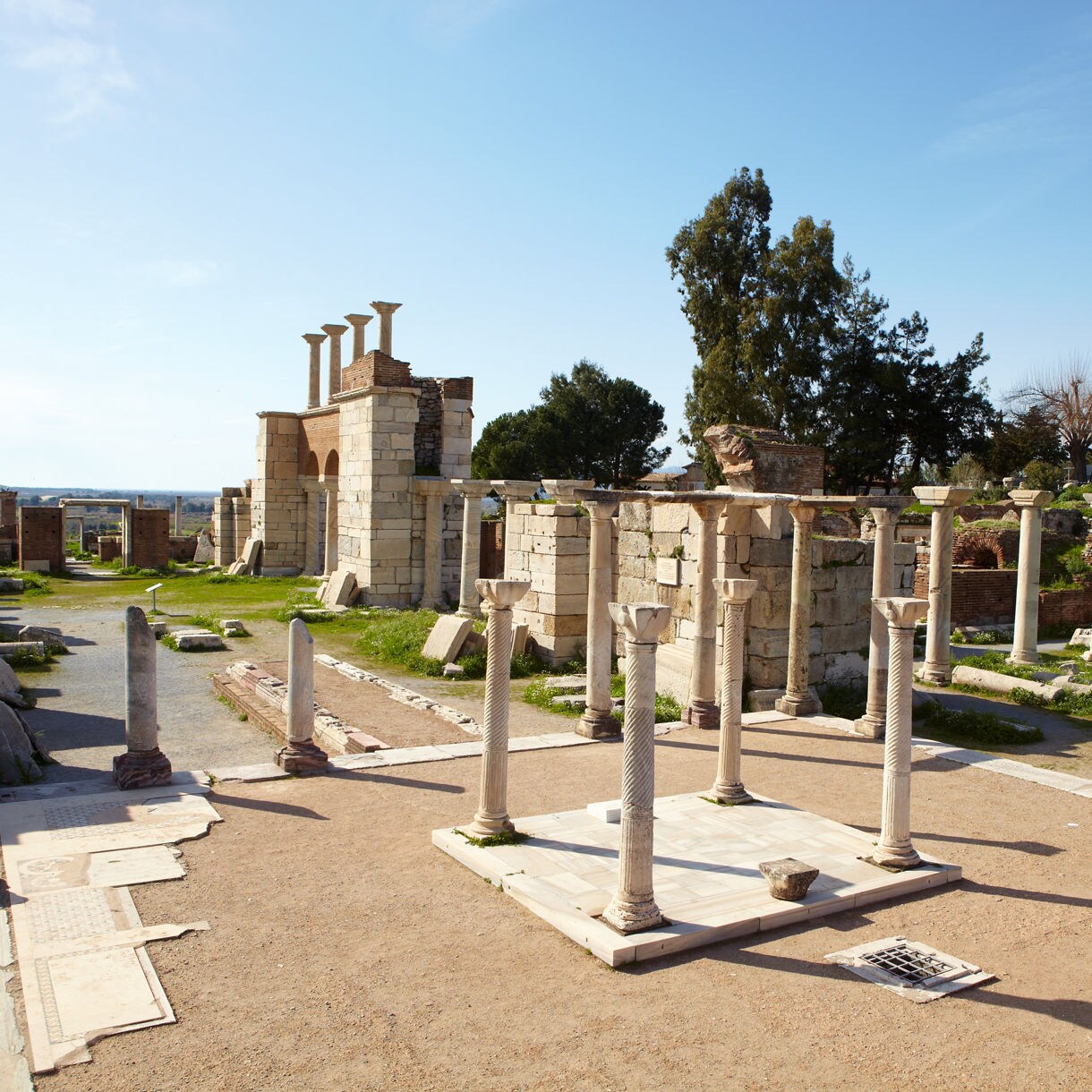 Ruins of the Basilica of St. John in Selçuk, Turkey, featuring scattered marble columns and stone walls under a bright blue sky with trees in the background.