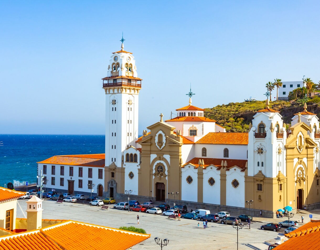 The Basílica de Candelaria in Tenerife, a white and beige church with red-tiled roofs and a tall bell tower overlooking the blue Atlantic Ocean.