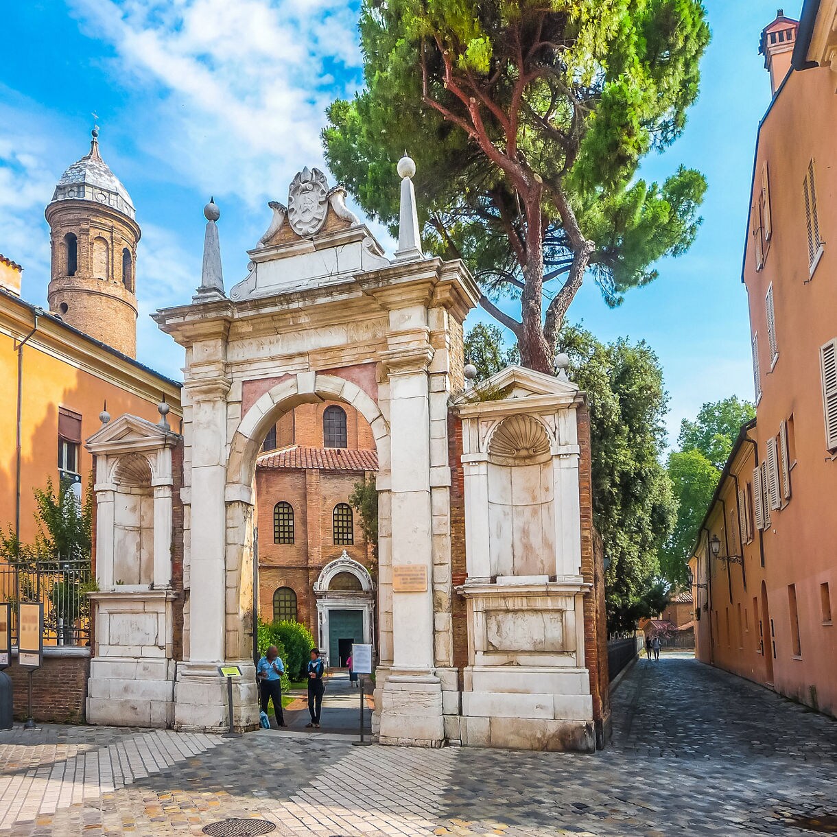 Step through the historic gateway to the Basilica of San Vitale, where centuries of art, architecture and history await in the heart of Ravenna. Alt text: Ornate stone entrance gate leading to the Basilica of San Vitale in Ravenna with a towering pine tree and historic brick church beyond.
