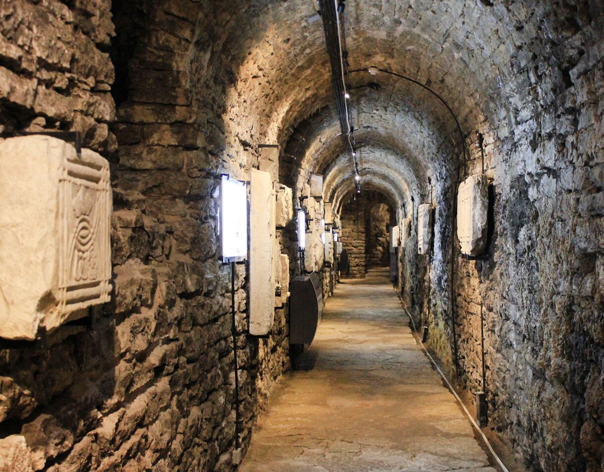 Stone-walled underground corridor of the Bastion Passages in Tallinn, Estonia, with arched ceilings, dim lighting and carved stone relics displayed along the walls.