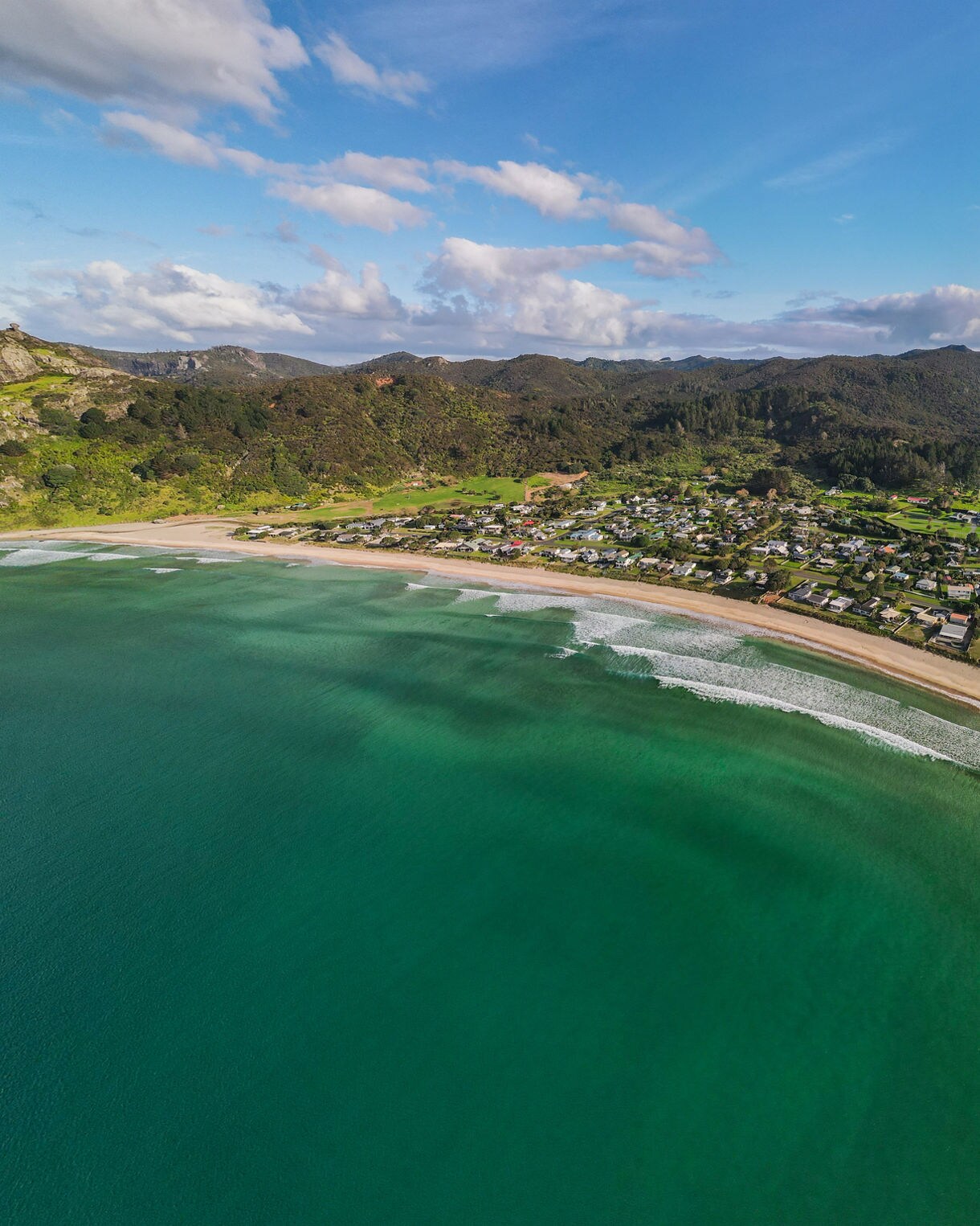 Aerial view of a long sandy beach with calm green ocean waves and a small coastal town backed by rolling hills in New Zealand’s Bay of Plenty.