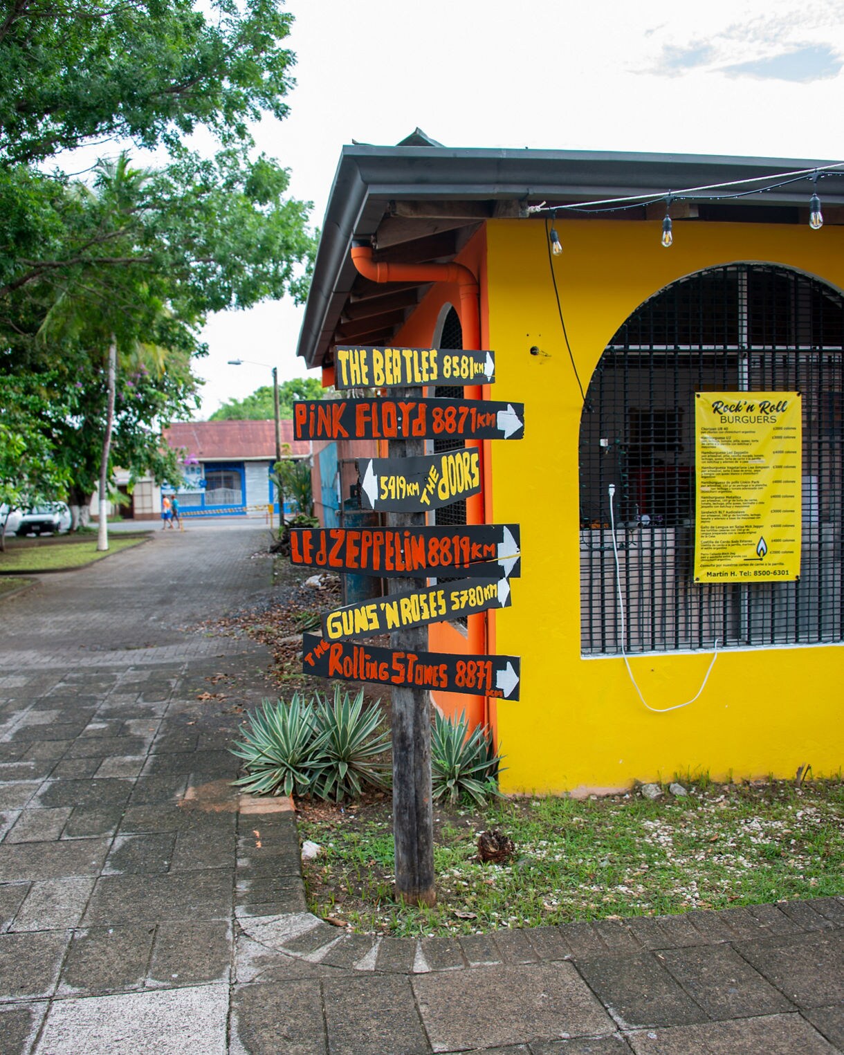 Wooden signpost with colorful arrows pointing to bands like The Beatles, Pink Floyd, Led Zeppelin and The Rolling Stones outside a bright yellow restaurant.