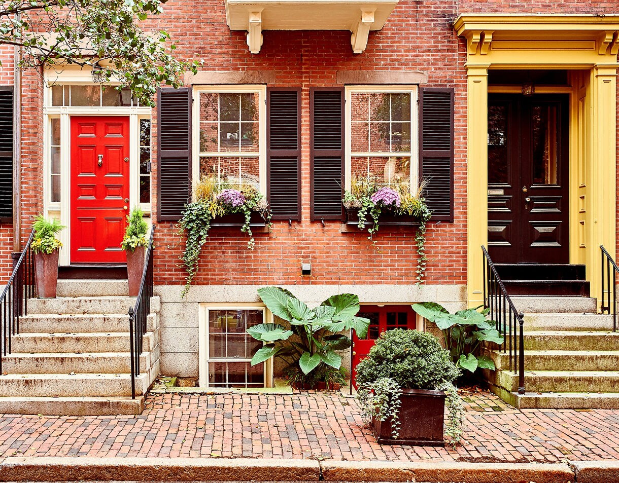 Brick townhouse façade in Beacon Hill with a bright red door, black shutters, window boxes filled with flowers and leafy plants along a cobblestone sidewalk.