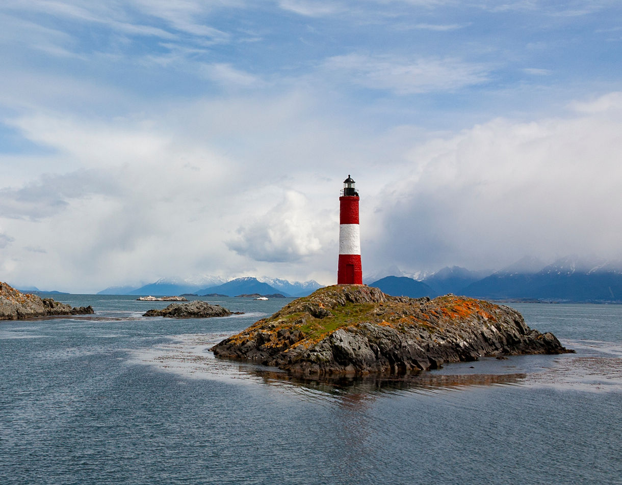 A red-and-white lighthouse standing on a rocky island in the Beagle Channel with small islets and snowy mountains in the background.