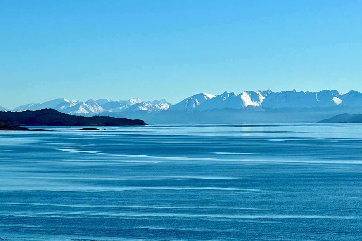 Wide view of the Beagle Channel with calm blue water, dark headlands and a distant range of snow-covered mountains under a clear sky.